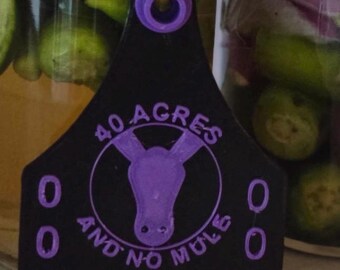 May include: Black tag with a purple mule graphic and the text "40 Acres and No Mule." The tag has a hole at the top and the numbers "001" on each side. The tag is in front of glass jars.