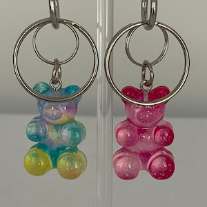 Puede incluir: Dos llaveros de ositos de gominola, uno azul y verde y otro rosa, colgando de llaveros plateados.