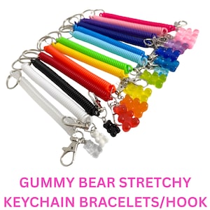 Puede incluir: Una colección de pulseras elásticas para llavero de colores con cierres metálicos y dijes de ositos de goma. Las pulseras vienen en varios colores, incluyendo blanco, negro, rojo, amarillo, verde, azul, morado, rosa y transparente. El texto "GUMMY BEAR STRETCHY KEYCHAIN BRACELETS/HOOK" se muestra en rosa.