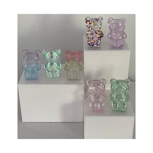 May include: A selection of small, translucent bear figurines in various colours, including blue, pink, green, and clear. Some bears are filled with colourful glitter. The bears are displayed on white platforms against a white background.