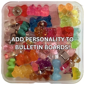 Pode incluir: Uma coleção de percevejos coloridos e translúcidos em forma de ursinhos de goma com pontas de metal, guardados num recipiente de plástico transparente. O texto ADD PERSONALITY TO BULLETIN BOARDS! está impresso em branco sobre os percevejos.