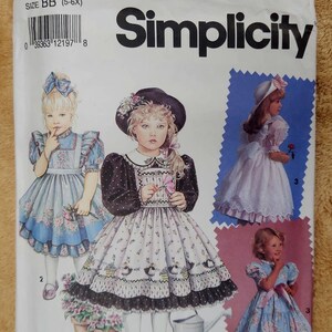 May include: A Simplicity 7699 sewing pattern envelope for size BB (5-6X) featuring illustrations of children wearing dresses with floral prints, puffed sleeves, and lace trim. The Daisy Kingdom brand logo is printed at the bottom of the white paper packet.