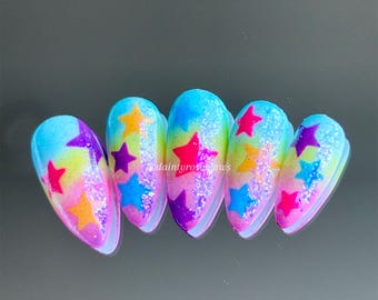 Puede incluir: Conjunto de seis uñas postizas coloridas con un diseño degradado arcoíris y purpurina. Cada uña presenta un diseño de estrella en varios colores, incluyendo rosa, azul, amarillo y morado.