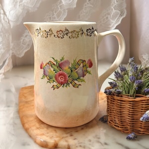 May include: A white ceramic pitcher with a gold trim and a floral design featuring thistles. The pitcher has a handle and a spout.