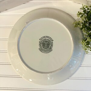 May include: A white ceramic plate with a gray rim and a decorative emblem in the center. The emblem features text and a detailed illustration. The plate is set against a white background with a wooden accent.