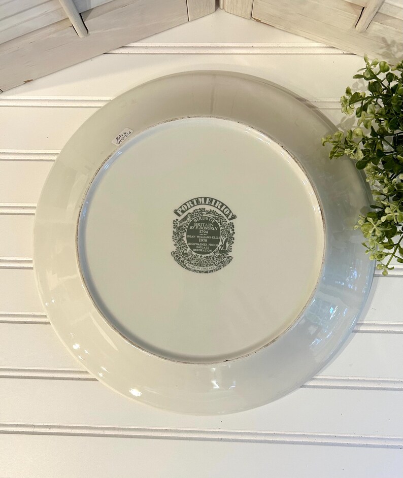 May include: A white ceramic plate with a gray rim and a decorative emblem in the center. The emblem features text and a detailed illustration. The plate is set against a white background with a wooden accent.