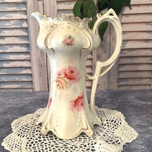 May include: Cream-colored porcelain pitcher with pink rose floral design and gold trim. The pitcher has a decorative handle and a scalloped rim. It is displayed on a cream-colored lace doily.