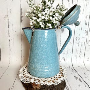 May include: A vintage-style, light blue enamelware pitcher with a matching lid, filled with white baby's breath flowers. The pitcher sits on a wooden slice with a white lace doily. The background is a white, weathered wood.