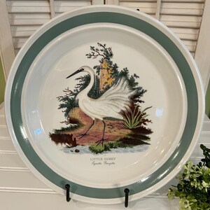 May include: A white ceramic plate with a teal green border and a detailed illustration of a Little Egret bird in a natural setting. The plate is displayed on a black metal stand, showcasing a vintage aesthetic.