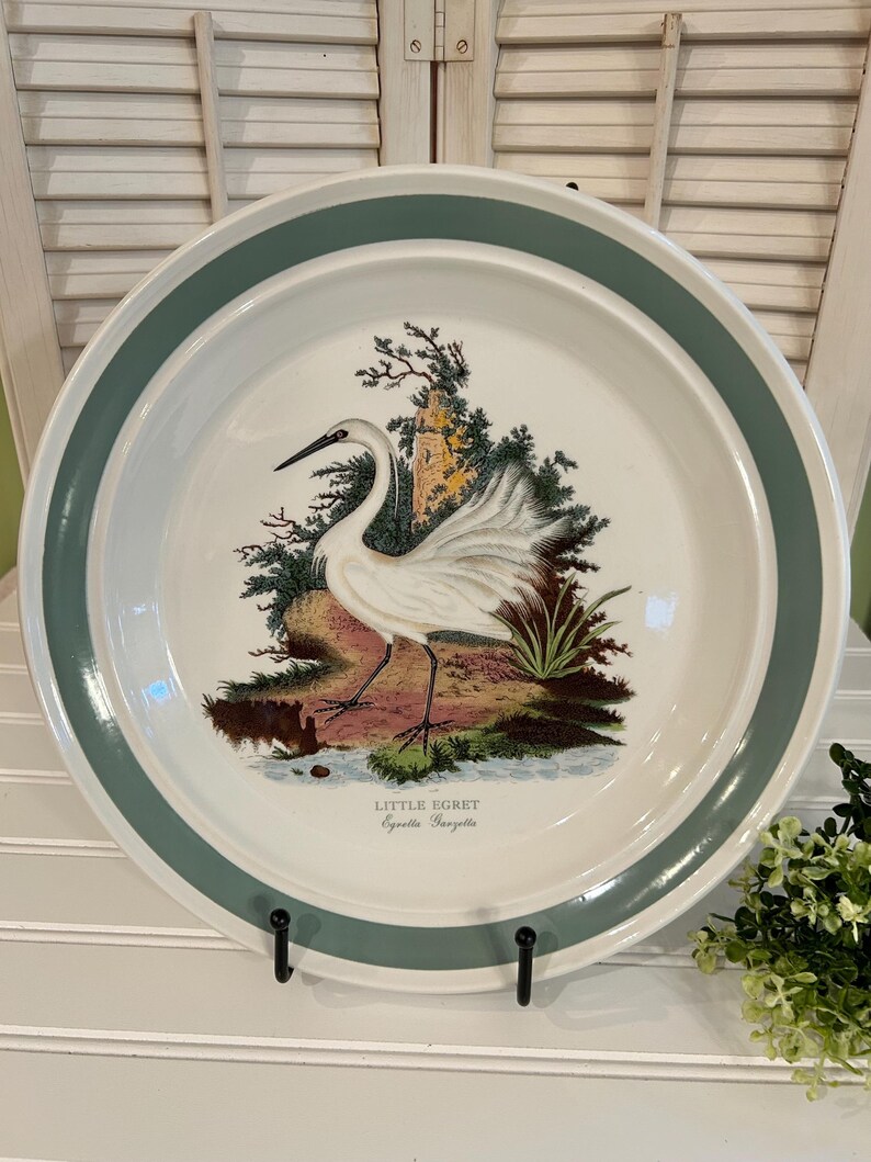 May include: A white ceramic plate with a teal green border and a detailed illustration of a Little Egret bird in a natural setting. The plate is displayed on a black metal stand, showcasing a vintage aesthetic.