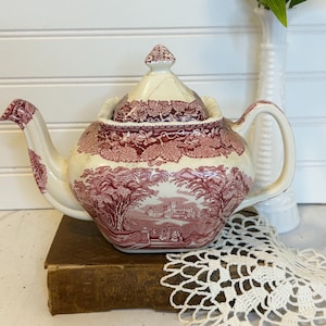 May include: Cream-colored teapot with a red scenic design. The teapot features a lid with a decorative finial and a curved handle. The design depicts a landscape with trees, buildings, and figures. The teapot is sitting on a vintage book.