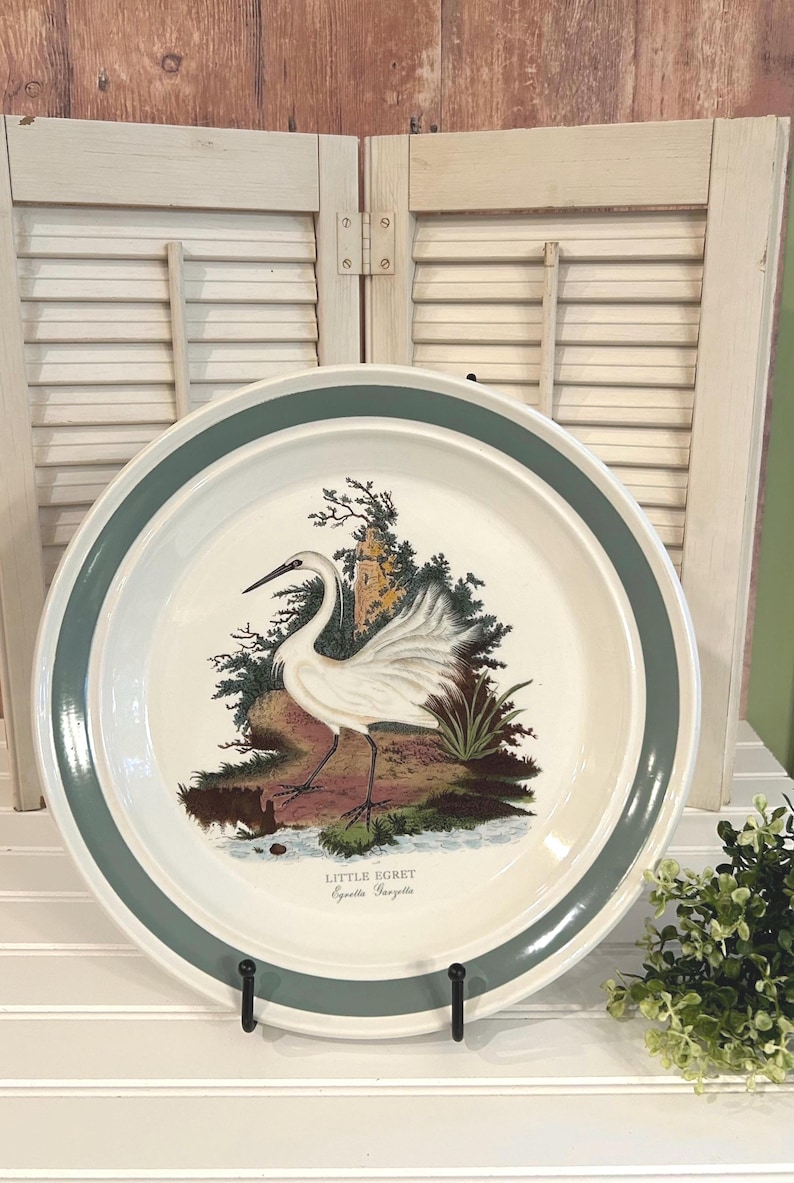 May include: A white ceramic plate with a teal green border. The plate features a detailed illustration of a Little Egret bird standing near a body of water. The bird is white with black legs and a long beak. The plate is displayed on a black metal stand.