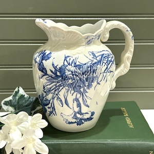 May include: A white ceramic pitcher with a blue floral design. The pitcher has a scalloped rim and a curved handle. The floral pattern is detailed and covers most of the pitcher's surface. The pitcher is sitting on a green book.
