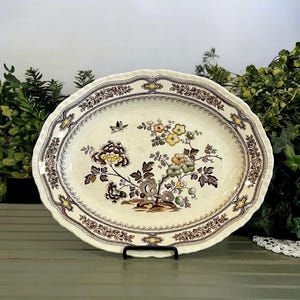 May include: Oval serving platter with a floral design in shades of brown, yellow, and green. The platter has a scalloped edge and a decorative border. The floral pattern includes a bird and stylized flowers.