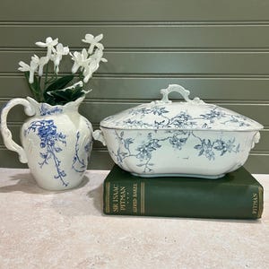May include: A white and blue floral pattern serving dish with a lid, a white pitcher with a blue floral design, and a green book. The serving dish and pitcher are on a light-colored surface.