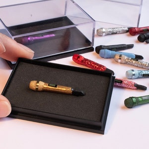 May include: A collection of miniature microphones in various colours and finishes, including gold, red, green, blue, and silver. Some microphones are covered in glitter. The microphones are displayed in a clear plastic box and on a white surface.