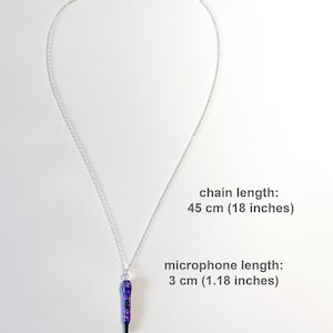 K&ouml;nnte beinhalten: Halskette mit silberner Kette und lila-schwarzem Mikrofonanh&auml;nger. Die Kette ist 45 cm lang, der Mikrofonanh&auml;nger 3 cm. Der Mikrofonanh&auml;nger hat lila Glitzerakzente.