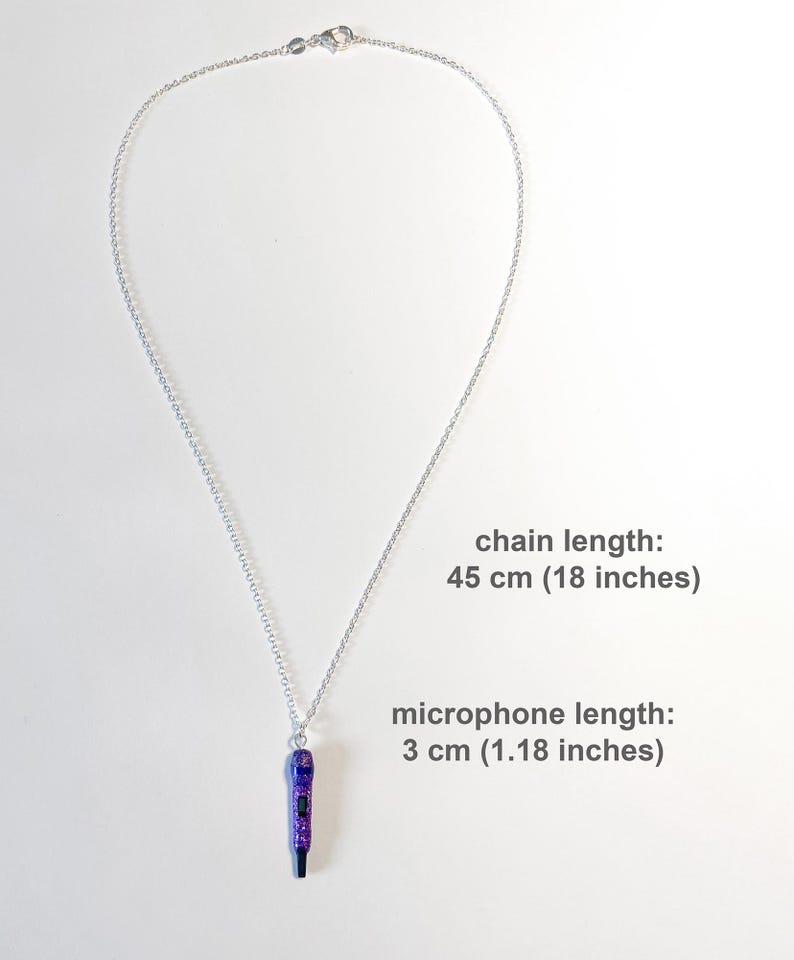 K&ouml;nnte beinhalten: Halskette mit silberner Kette und lila-schwarzem Mikrofonanh&auml;nger. Die Kette ist 45 cm lang, der Mikrofonanh&auml;nger 3 cm. Der Mikrofonanh&auml;nger hat lila Glitzerakzente.