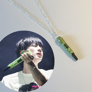 Puede incluir: Un collar de cadena plateada con un colgante de micrófono verde. El micrófono está cubierto de purpurina y tiene una punta negra. La imagen también incluye una foto circular de una persona sosteniendo un micrófono similar.