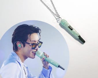 Pode incluir: Um colar de corrente prateada com um pendente em forma de microfone verde claro. O pendente do microfone é coberto de glitter e tem uma ponta preta. O colar é exibido em um fundo branco.