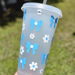 May include: A clear plastic tumbler with a blue and white design. The tumbler has a straw and is decorated with blue bows and white flowers.