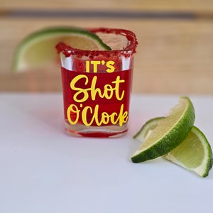 May include: A red shot glass with a sugared rim and the text "It's Shot O'Clock" in yellow lettering.  A lime wedge is behind the glass and three lime wedges are in front of the glass.