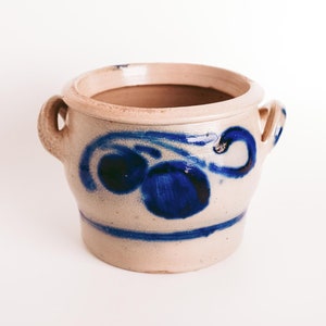May include: A small, white ceramic pot with blue painted floral designs. The pot has two handles on the sides.