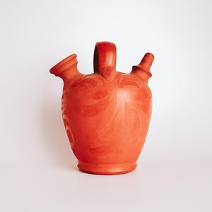 May include: A terracotta pitcher with a unique design and two spouts. The pitcher is reddish-brown in colour and has a textured surface.