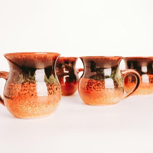 May include: Four brown ceramic mugs with green and orange glaze. The mugs have a rounded body and a handle.