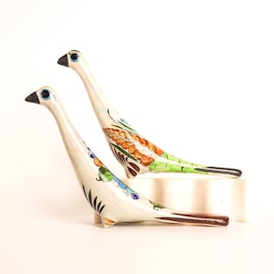 May include: Two ceramic bird ornaments with elongated necks and painted details. The birds feature black beaks, blue eyes, and colourful patterns on their bodies, including orange, green, and blue accents. The ornaments are displayed on a white surface.