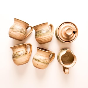 May include: Set of six handmade ceramic mugs and a creamer in a rustic style with brown, beige, and green glaze.