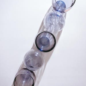 Pu&ograve; includere: Cinque bicchieri di vetro trasparente con una leggera sfumatura blu, disposti verticalmente in fila. I bicchieri sono vuoti e sembrano essere fatti di vetro spesso.