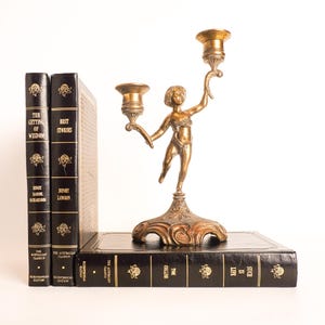 May include: A brass candelabra featuring a central figure of a child holding two candle cups, standing on a decorative base. The candelabra is positioned on a black book, next to two other black books with gold lettering and floral accents.