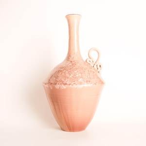 May include: A peach-coloured ceramic vase with a slender neck and a rounded body. The vase has a textured, crystalline pattern around the shoulder and a looped handle. The base is flared. The vase is set against a white background.