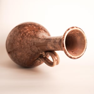 May include: A brown ceramic jug with a round body, a handle, and a flared spout. The jug has a textured surface with a speckled pattern. The handle is a loop shape. The jug is likely used for pouring liquids.