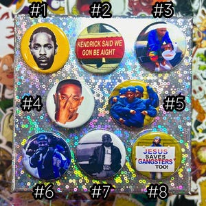 以下が含まれることがあります： 8つの丸いピンバッジのコレクション。バッジには、ラッパーの画像と「Kendrick Said We Gon Be Aight」や「Jesus Saves Gangsters Too！」などのテキストが表示されています。バッジは、キラキラ光る虹色の表面に配置されています。