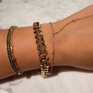 May include: A collection of gold bracelets and a delicate necklace. The bracelets include a thick, chain-link style, a tennis bracelet with green stones, and a dainty necklace with small diamonds.
