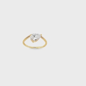 May include: A gold ring featuring a single pear-shaped clear gemstone set in a prong setting. The band is thin and polished, showcasing a simple design that highlights the faceted cut of the stone, which reflects light from its pointed top.