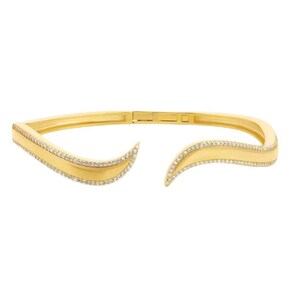 May include: A gold-toned open cuff bracelet featuring a curved, wave-like design. The edges of the bracelet are lined with small, clear, sparkling stones, while the center of the band remains a smooth, polished gold-colored metal finish.