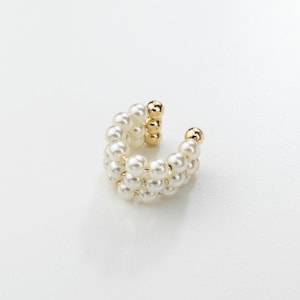 Peut inclure: Une boucle d'oreille ear cuff dorée composée de trois rangées parallèles de perles blanches sphériques. Le design circulaire est ouvert sur un côté, révélant une structure métallique dorée reliant les perles pour un aspect texturé.
