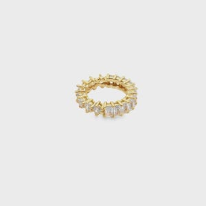 May include: A gold-toned ring featuring a continuous band of alternating rectangular baguette-cut and round-cut clear stones set in a prong setting. The metal band has a polished finish and the stones are arranged in a repeating pattern around the ring.