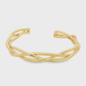 May include: A gold-toned cuff bracelet featuring a polished finish and a multi-strand design. The structure consists of several thin, smooth metal wires that are woven together in a continuous, overlapping pattern to form a circular open band.