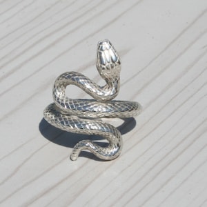 May include: A silver snake ring with a textured, scale-like pattern. The snake is coiled, with its head raised, creating a detailed piece of jewellery. The ring is displayed on a light-coloured wooden surface.