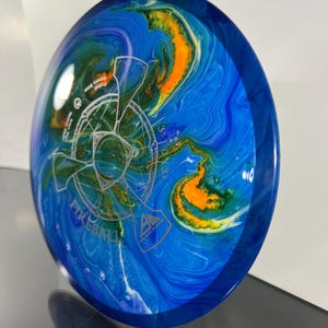 May include: Axiom Discs Fireball disc golf disc. The disc features a blue, orange, and green marbled design with a white graphic and the word "Fireball". The disc has a dark blue rim.