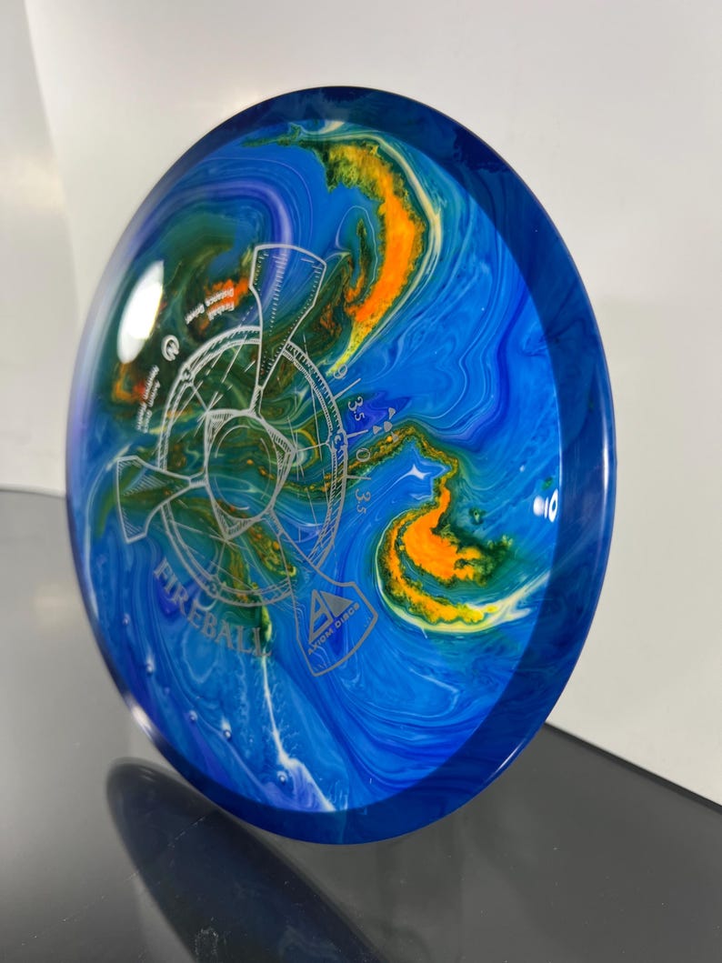 May include: Axiom Discs Fireball disc golf disc. The disc features a blue, orange, and green marbled design with a white graphic and the word "Fireball". The disc has a dark blue rim.