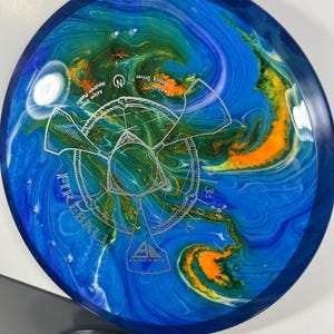 May include: Axiom Discs Fireball distance driver disc. The disc features a blue, green, and orange marbled design with the word "Fireball" and a graphic in the center. The disc is made of Neutron plastic.