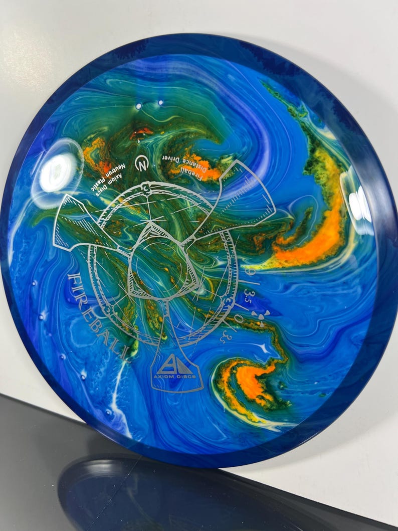 May include: Axiom Discs Fireball distance driver disc. The disc features a blue, green, and orange marbled design with the word "Fireball" and a graphic in the center. The disc is made of Neutron plastic.