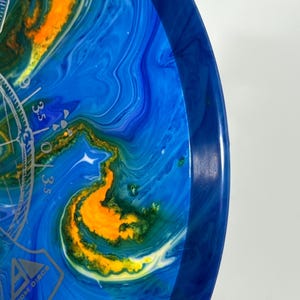 May include: Axiom Discs disc golf disc with a blue, yellow, and green marbled design. The disc has a blue rim and a silver graphic with the text "Axiom Discs". The disc has a diameter of approximately 8.5 inches.