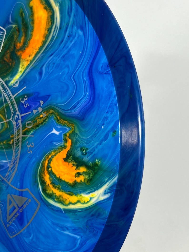 May include: Axiom Discs disc golf disc with a blue, yellow, and green marbled design. The disc has a blue rim and a silver graphic with the text "Axiom Discs". The disc has a diameter of approximately 8.5 inches.