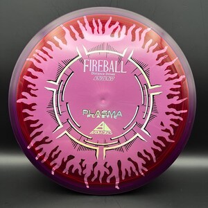 May include: A pink and red Fireball distance driver disc with the text "Fireball" and "Plasma Plastic" printed on it. The disc has a unique design with a central circular pattern and flame-like edges. The disc is made by Axiom Discs.